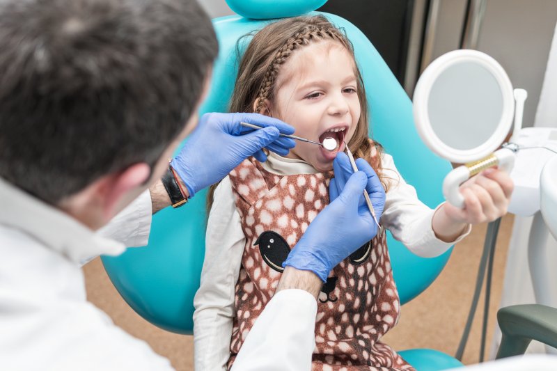 a dentist checking a little girl’s mouth while she holds a mirror